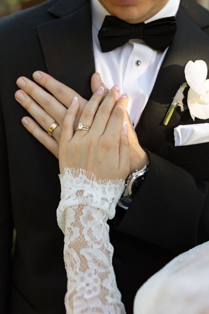 Close-up of bride's lace-sleeved hand with wedding ring resting on groom's tuxedo, white boutonniere visible