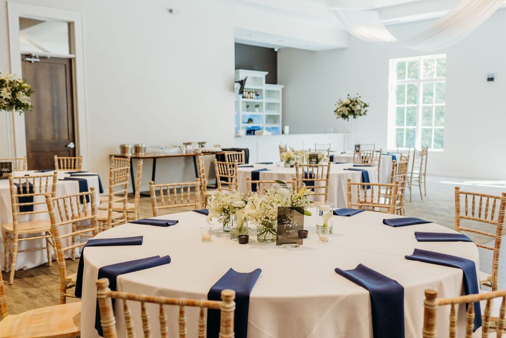 Wedding reception table setup with white linens, navy napkins, gold chiavari chairs, and white floral centerpieces in a bright venue space