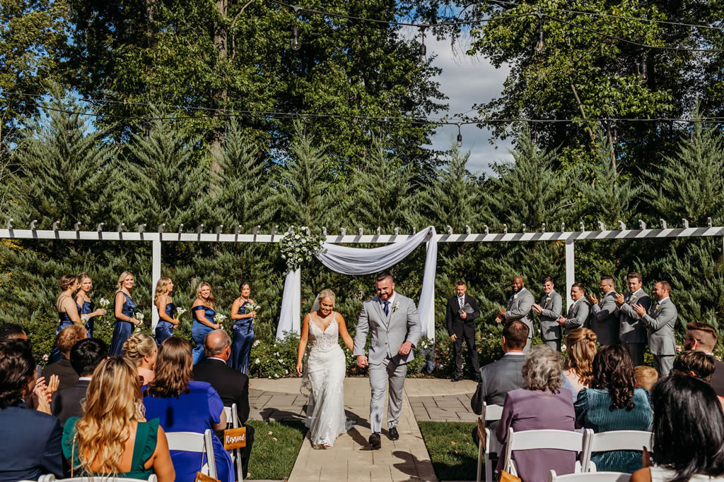 Outdoor wedding ceremony with bride and groom walking down aisle under white pergola, bridesmaids in navy and groomsmen in grey, guests seated on white chairs