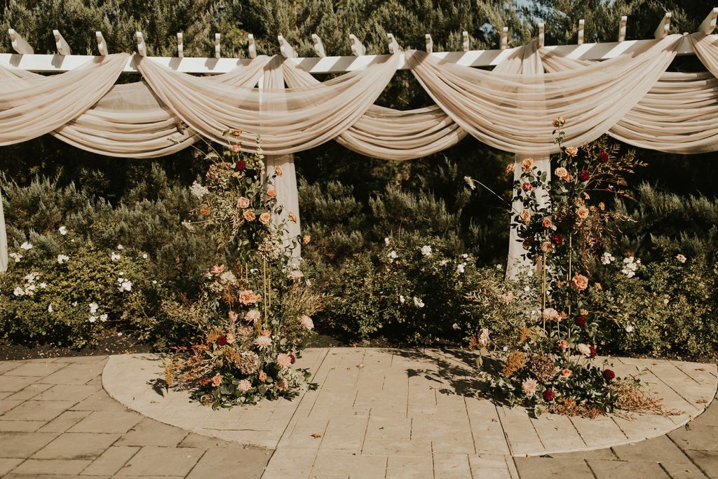 Outdoor wedding ceremony setup with draped white fabric canopy and tall floral arrangements featuring peach and burgundy roses"