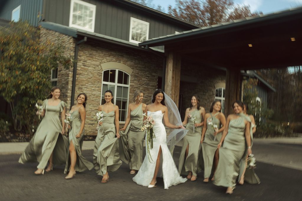 Bride and bridesmaids walking and laughing outside stone venue building, wearing white wedding gown and sage green bridesmaid dresses