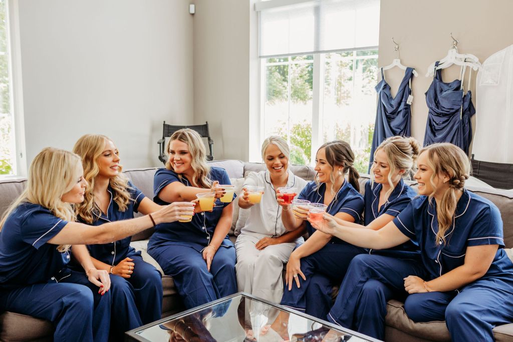 Bride in white robe and bridesmaids in navy pajamas toasting with colorful drinks in bridal suite, bridesmaid dresses hanging in background