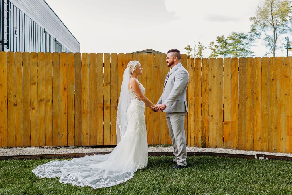 Bride in lace wedding gown and groom in light grey suit holding hands against wooden fence, bride's veil trailing on grass