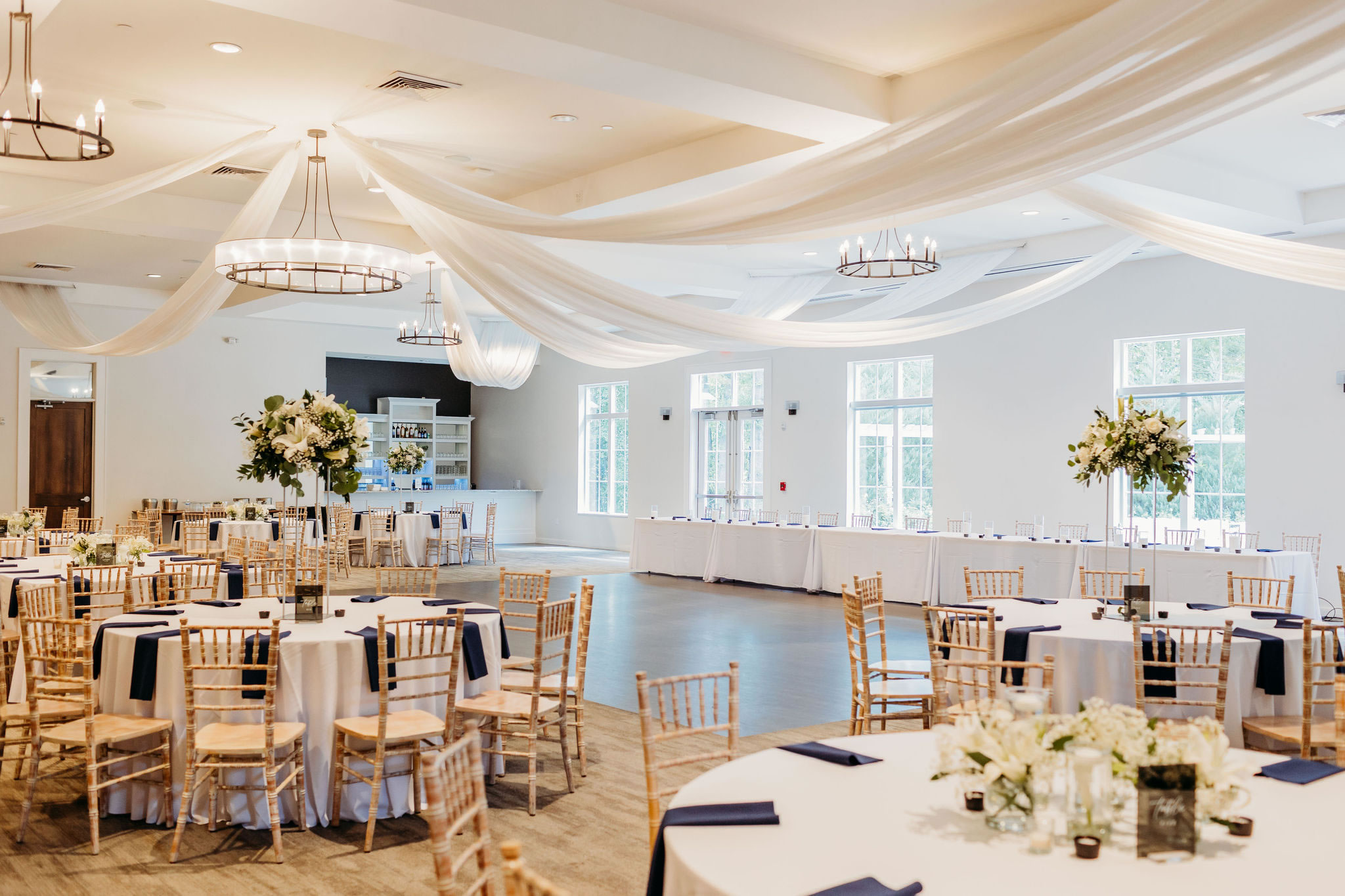 A bright and airy wedding reception hall with white draped ceilings, floral centerpieces, and wooden chairs.