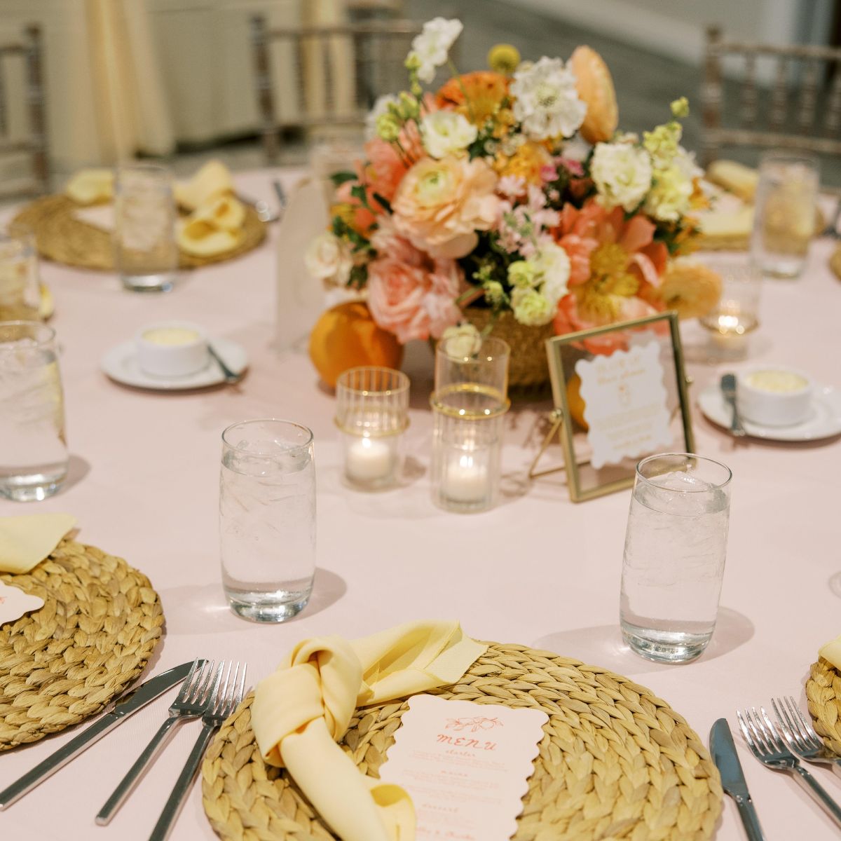 Corporate banquet table at The Estate at New Albany with woven chargers, yellow napkins, pastel floral centerpiece, candles, and printed menu cards.