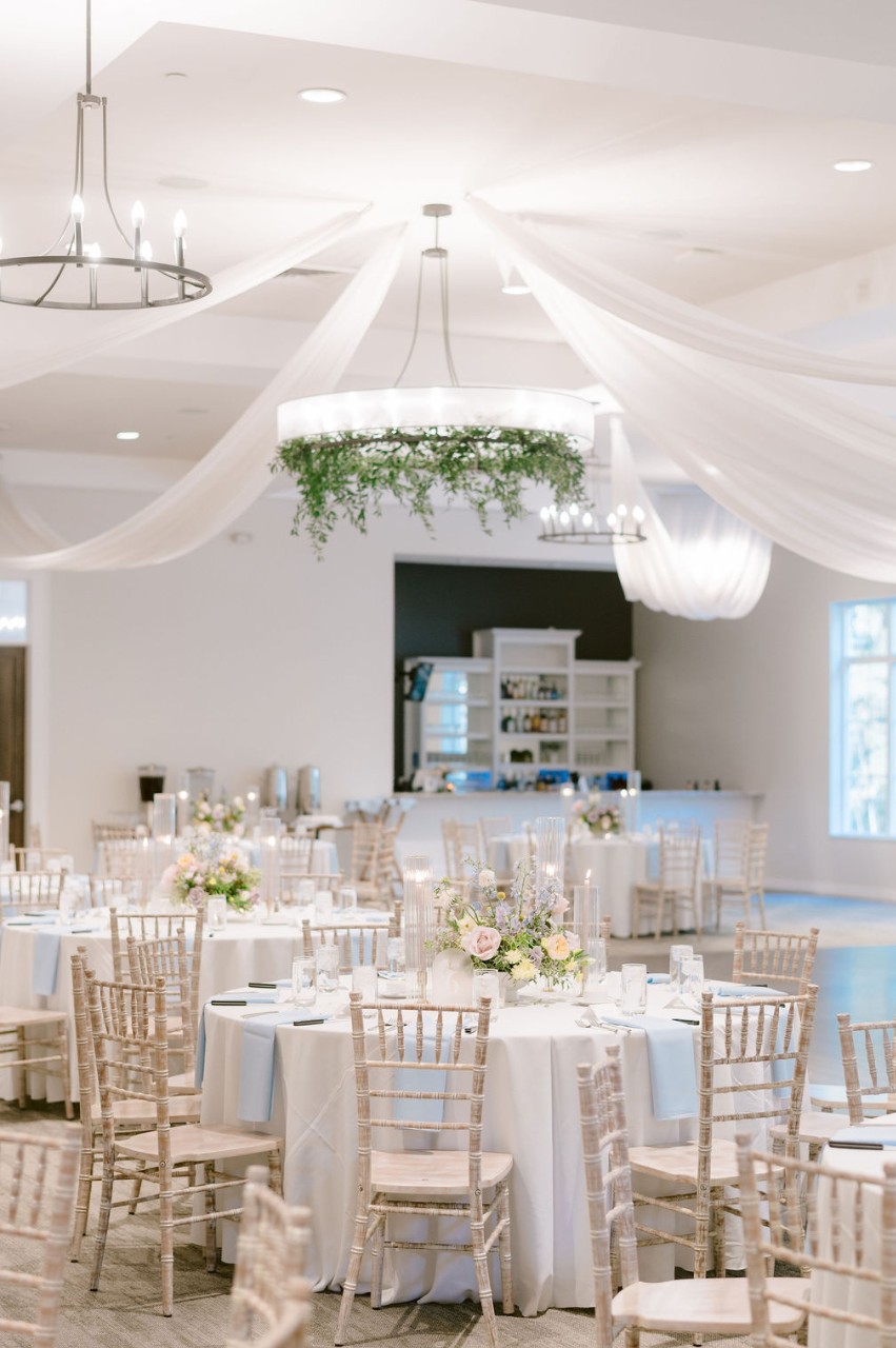 Wedding reception setup in the Great Hall at The Estate at New Albany, with round tables, pastel floral centerpieces, chiavari chairs, and draped ceiling décor.