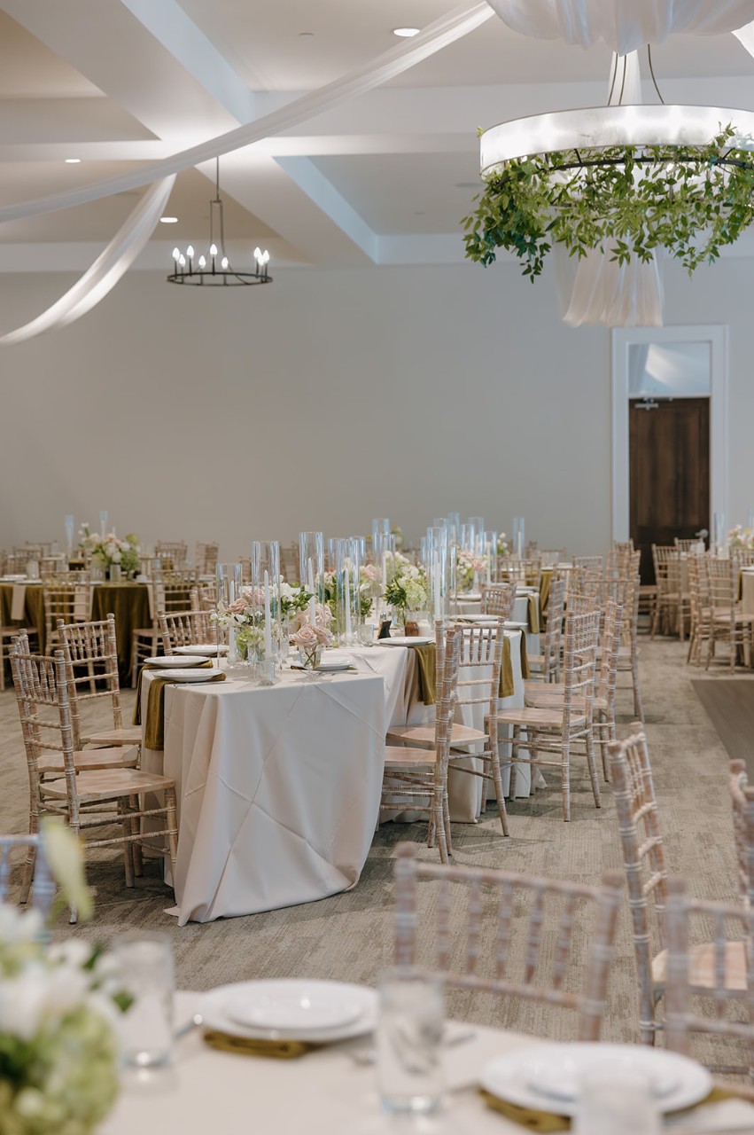 Wedding reception in the Great Hall at The Estate at New Albany, featuring banquet-style tables with white linens, greenery accents, and tall glass candle holders.