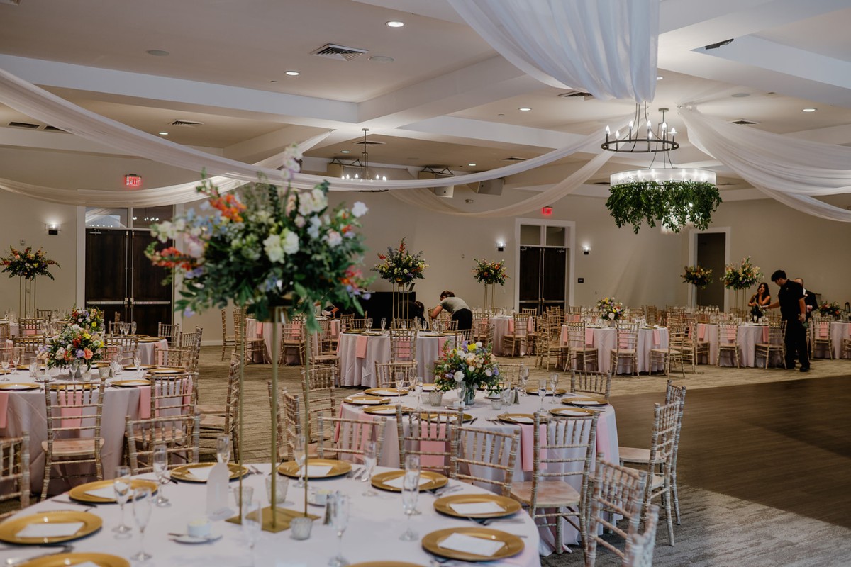 Wedding reception in the Great Hall at The Estate at New Albany, featuring round tables with gold chargers, pink linens, tall floral centerpieces, and draped ceiling décor.