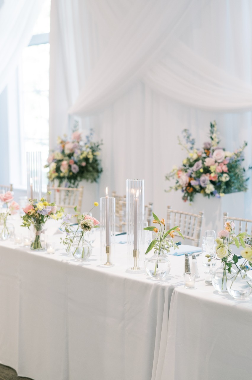 Wedding head table at The Estate at New Albany featuring pastel flower centerpieces, tall glass candleholders, chiavari chairs, and a draped fabric backdrop for a ballroom reception.