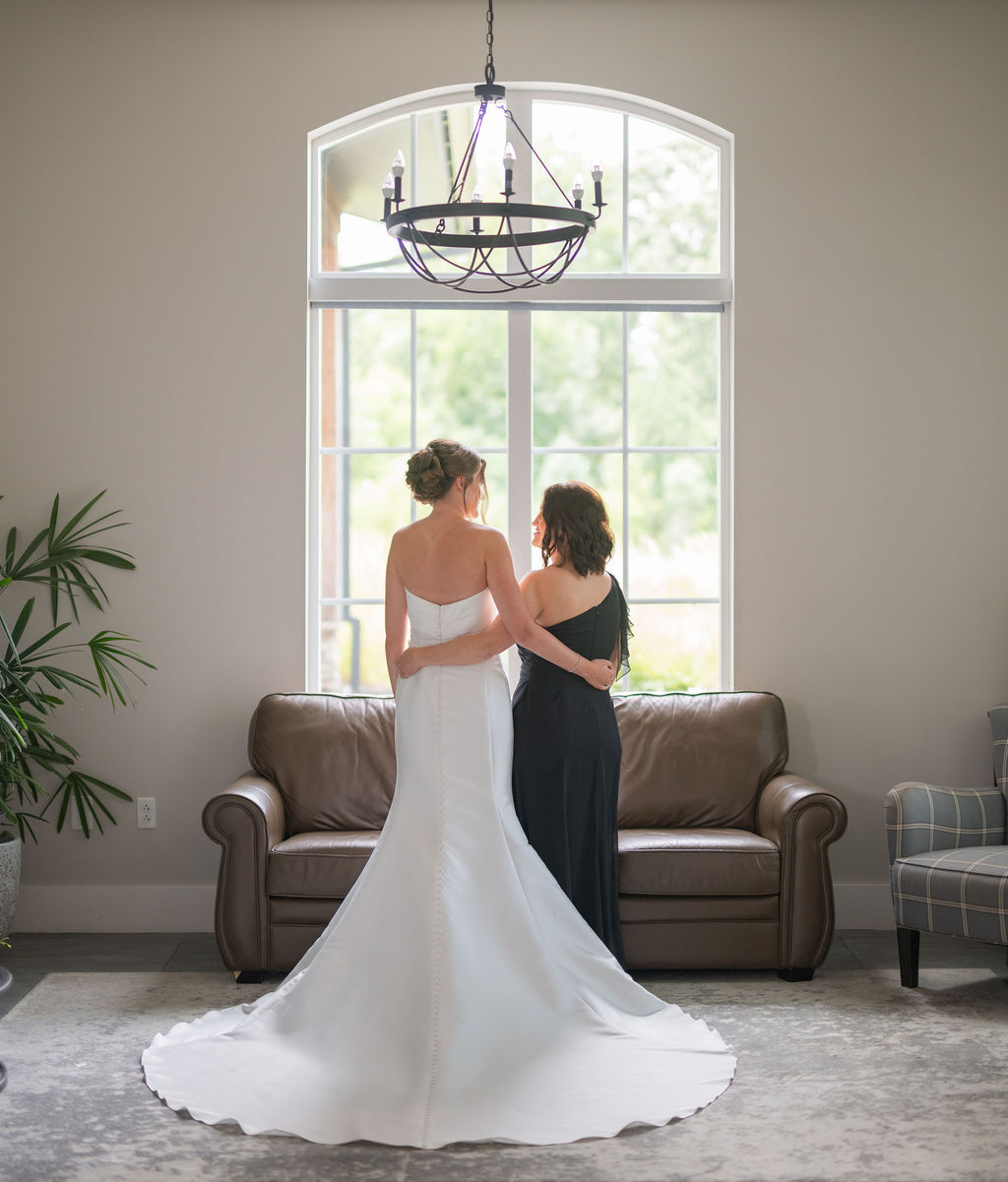 Bride in white and her mother in black dress embracing facing window